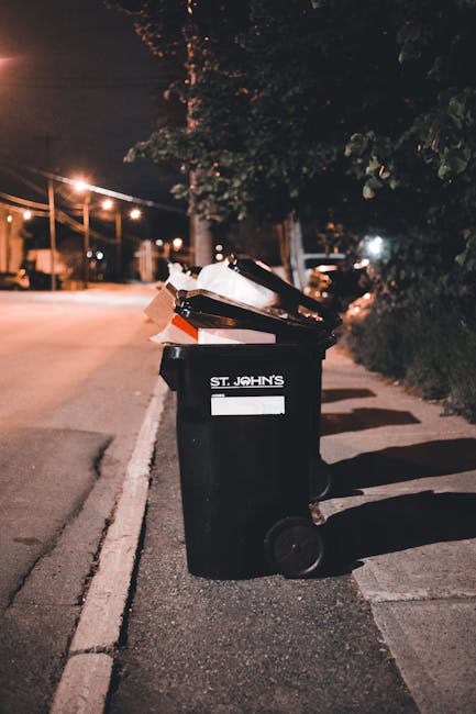 A dark green wheeled rubbish bin with a white label reading 'St. John's' is positioned on the edge of a pavement alongside a quiet street at night. The bin is filled with various discarded waste items, including a black plastic tray, paper, cardboard, and possibly some plastic packaging, which are spilling over the top. The surface of the bin appears slightly weathered with a matte finish. The background shows a row of illuminated streetlights casting a warm glow over the tarmac road and bordering foliage, while a few distant cars are faintly visible. The pavement is made of concrete slabs, and the location appears to be a residential or semi-urban area with trees and shrubs along the roadside. Nearby patches of shadow cast by the streetlights create a subdued atmosphere, consistent with routine waste disposal, and the scene subtly reflects private or independent rubbish handling suitable for professional waste collection services like Waste Collection Camden.