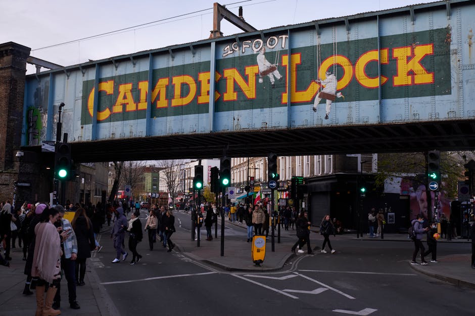 A busy street scene in Camden Town featuring a large, weathered metal railway bridge overhead, painted with the words 'Camden Lock' in bold yellow and red lettering. The bridge spans across the intersection, supporting electrical wires and structural beams. Below, pedestrians cross the road on a mixture of paved sidewalks and crosswalks, with some waiting at traffic lights and others walking in different directions. The crowd includes people dressed in casual coats, scarves, and backpacks, reflecting a lively urban atmosphere. On the left side, a traffic light displays a green signal, guiding vehicles and cyclists along the street. The environment includes storefronts and buildings lining the background, with some shopfronts partially visible and illuminated shop signs. The overall lighting suggests late afternoon or early evening, with a soft, subdued natural light casting gentle shadows. The image subtly relates to alternative waste handling by illustrating a typical urban setting where waste collection services like Waste Collection Camden may operate to manage rubbish generated by the busy community, especially in a vibrant area like Camden Town.
