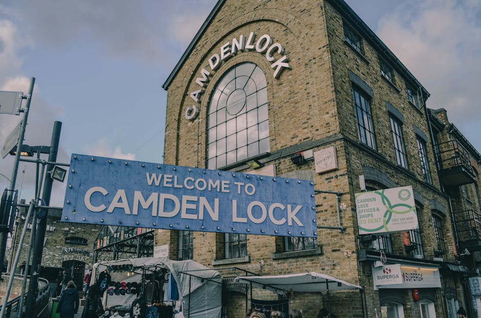 A busy street scene in Camden Town featuring a large, weathered metal railway bridge overhead, painted with the words 'Camden Lock' in bold yellow and red lettering. The bridge spans across the intersection, supporting electrical wires and structural beams. Below, pedestrians cross the road on a mixture of paved sidewalks and crosswalks, with some waiting at traffic lights and others walking in different directions. The crowd includes people dressed in casual coats, scarves, and backpacks, reflecting a lively urban atmosphere. On the left side, a traffic light displays a green signal, guiding vehicles and cyclists along the street. The environment includes storefronts and buildings lining the background, with some shopfronts partially visible and illuminated shop signs. The overall lighting suggests late afternoon or early evening, with a soft, subdued natural light casting gentle shadows. The image subtly relates to alternative waste handling by illustrating a typical urban setting where waste collection services like Waste Collection Camden may operate to manage rubbish generated by the busy community, especially in a vibrant area like Camden Town.