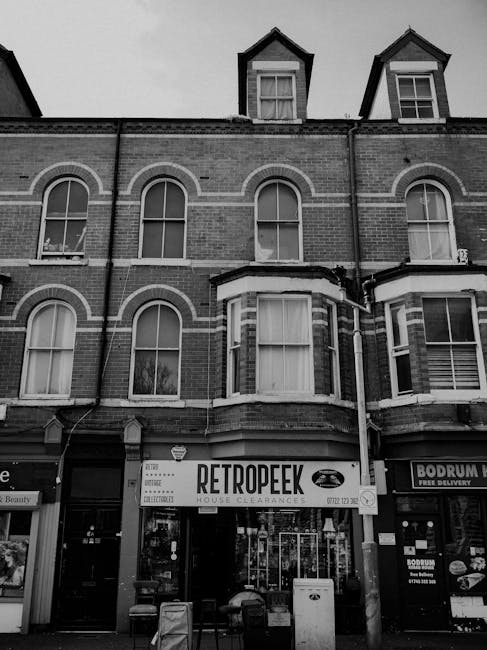 The image depicts the façade of a multi-storey brick building with three visible floors, featuring a traditional Victorian-style design with arched window frames, some with visible curtains behind the glass. The building’s exterior walls are brick with light-colored mortar, and the windows on the upper floors are sash windows, some open slightly and others closed. A prominent bay window extends from the ground and first floors, with a flat roof and decorative brickwork. On the ground floor, there are commercial shopfronts, including a retail store named 'RetropEEK' specializing in house clearances, with a large sign displaying the store name and a stylized vintage car logo, as well as another shop next door offering food delivery services, evident from the signage and windows. The shopfronts are partially obscured by waste bins and small barricades placed on the pavement outside, possibly indicating recent rubbish collection or preparation for waste removal. The scene is captured in black and white, emphasizing textures and contrasts of the brickwork, windows, and urban environment, reflecting typical private waste handling activities in a city setting associated with rubbish removal services.