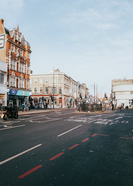 A street scene in an urban area with a wide road carrying multiple lanes of traffic, including a bus lane marked with white letters and red dashed lines. On the left, there are several mid-rise buildings with ornate architectural details, brickwork, and large windows, some with shops on the ground floor. Pedestrians are visible walking along the pavement, crossing at designated crosswalks near bus stops with shelter structures. In the background, additional buildings and street furniture such as lampposts and traffic signals can be seen, under a clear sky with soft, natural daylight. The environment appears to be a busy city centre, and the scene may relate to local waste management including rubbish collection or private waste disposal services, supported by visible activity near the curb and the presence of waste collection professionals possibly engaged in rubbish removal operations noted by waste collection Camden, subtly aligning with private or alternative waste handling methods.