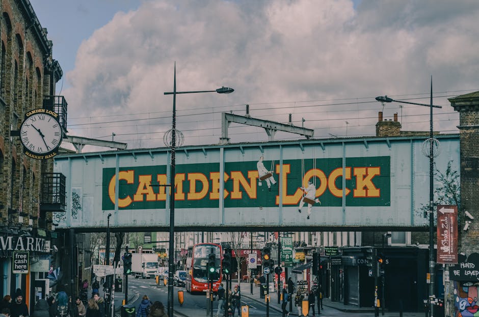 The image displays a busy urban street scene beneath a large, vintage-style train bridge painted with the words 'Camden Lock' in bold yellow letters on a dark green background. The bridge structure is made of weathered metal, with visible rivets and a slightly distressed finish. On the left side, part of an old brick building with a clock mounted on the facade is visible, showing the time as approximately 12:08. Streetlights with elongated arms extend above the street, casting a soft light across the scene. Below the bridge, numerous pedestrians are seen walking along the pavement, some gathered near shops and street vendors. A red double-decker bus is present in the middle distance, contributing to the lively atmosphere of the area. The street is filled with various vehicles, including a white van and smaller cars, while traffic signals and street signage guide the flow of traffic. The environment appears to be a vibrant, urban location with a mix of commercial activity and public transportation, consistent with an area where private waste removal and alternative rubbish collection services, such as those provided by Waste Collection Camden, might operate in support of maintaining cleanliness and waste management in a busy marketplace setting.