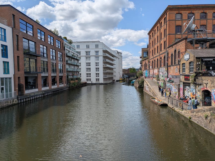 A canal scene in an urban environment with modern and historic buildings lining the waterway. On the left, there are contemporary multi-storey residential apartment buildings constructed with red brick and glass, featuring large windows and small balconies, set close to the canal edge. On the right, there is an older red-brick industrial building with arched windows, graffiti-covered walls, and external fire escape stairs, indicating a creative or mixed-use area. The canal water is calm, reflecting the partly cloudy sky above, with a slight ripple on the surface. A group of people stand near the canal on the right side, some observing the view and others engaged in conversation. A small wooden boat is moored along the edge of the historic building, partly obscured by a metal railing. The environment appears to be a lively part of the city that may involve private waste handling and on-site clearance activities, consistent with alternative rubbish collection services such as those provided by Waste Collection Camden, situated in an area with a mixture of residential, commercial, and artistic spaces.