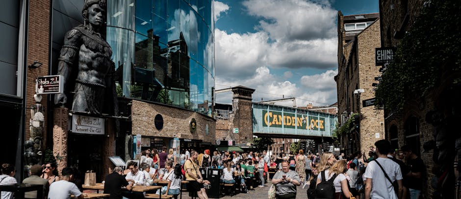 The image displays a busy urban street scene beneath a large, vintage-style train bridge painted with the words 'Camden Lock' in bold yellow letters on a dark green background. The bridge structure is made of weathered metal, with visible rivets and a slightly distressed finish. On the left side, part of an old brick building with a clock mounted on the facade is visible, showing the time as approximately 12:08. Streetlights with elongated arms extend above the street, casting a soft light across the scene. Below the bridge, numerous pedestrians are seen walking along the pavement, some gathered near shops and street vendors. A red double-decker bus is present in the middle distance, contributing to the lively atmosphere of the area. The street is filled with various vehicles, including a white van and smaller cars, while traffic signals and street signage guide the flow of traffic. The environment appears to be a vibrant, urban location with a mix of commercial activity and public transportation, consistent with an area where private waste removal and alternative rubbish collection services, such as those provided by Waste Collection Camden, might operate in support of maintaining cleanliness and waste management in a busy marketplace setting.