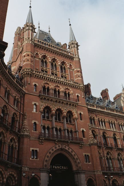 A tall, historic red-brick building with ornate architectural details, featuring multiple arched windows, decorative stonework, and prominent spires at the top. The structure has a medieval or Gothic Revival aesthetic, with a large central archway at the ground level leading into the entrance. The sky above is overcast with grey clouds, casting diffuse lighting across the facade. The building appears to be part of a well-known landmark, possibly a train station or civic building, situated in an urban environment. While the image does not explicitly show rubbish or waste, this type of structure is often associated with urban waste collection or hosting facilities. The surroundings in the lower part include parts of adjacent buildings and street-level features, which may relate to local waste management or private clearance services, such as Waste Collection Camden, operating in the vicinity.