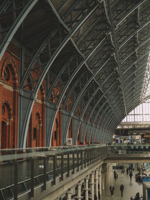 A spacious interior view of St Pancras station featuring an arched, modern steel and glass roof structure with curved steel beams painted dark grey, supporting large glass panels that allow natural light to illuminate the station. The lower level includes a wide walkway with a metal railing along the edge, overlooking the busy concourse below where passengers are walking and standing. On the left side, part of a traditional red brick wall with arched windows is visible behind the steel framework, contrasting with the contemporary architectural elements. The overall atmosphere is bright, clean, and industrial, highlighting the station’s blend of historic and modern design, with an emphasis on the structural details of the roof and the bustling activity of passengers below, consistent with the context of handling different types of waste removal and site clearance in large transit hubs.